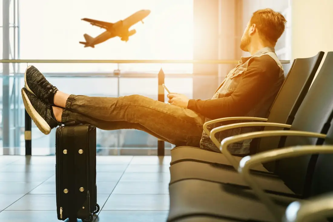 A business traveler waits at an airport gate with a carry-on as a jet takes off outside the window